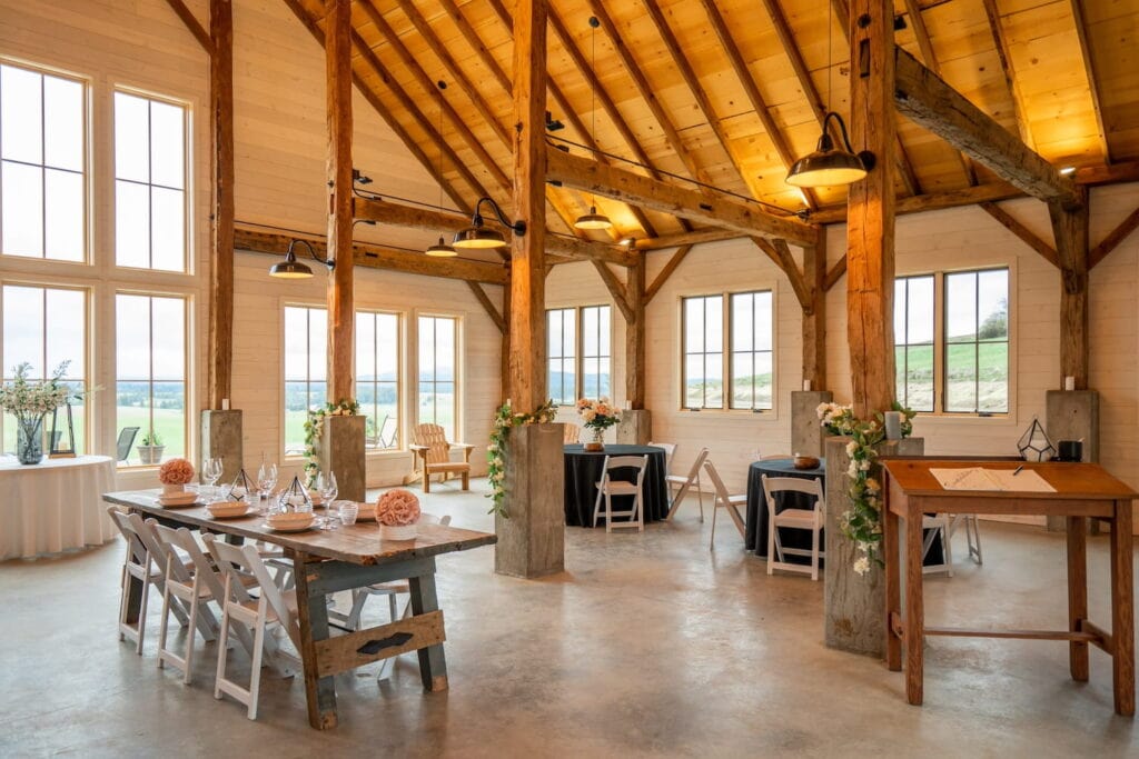 Simple barn reception with wooden tables, white chairs and scattered floral arrangements beneath wooden beams and illuminated by large paned windows